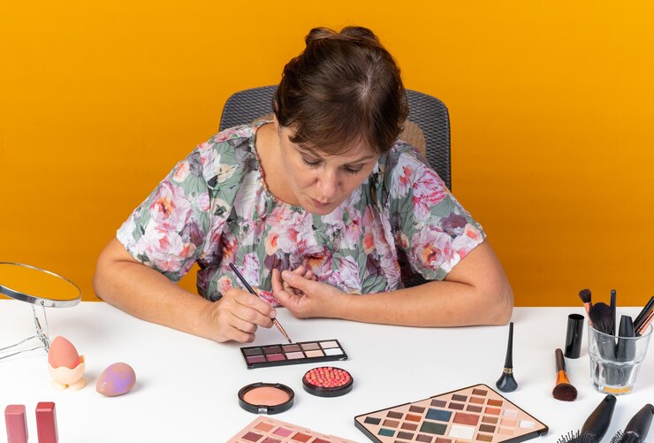 Foto gratuito segura mujer caucásica adulta sentada a la mesa con herramientas de maquillaje sosteniendo pincel de maquillaje y mirando la paleta de sombra de ojos aislada en la pared naranja con espacio de copia