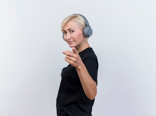 Segura mujer eslava rubia de mediana edad con auriculares de pie en la vista de perfil mirando y apuntando a la cámara aislada sobre fondo blanco con espacio de copia