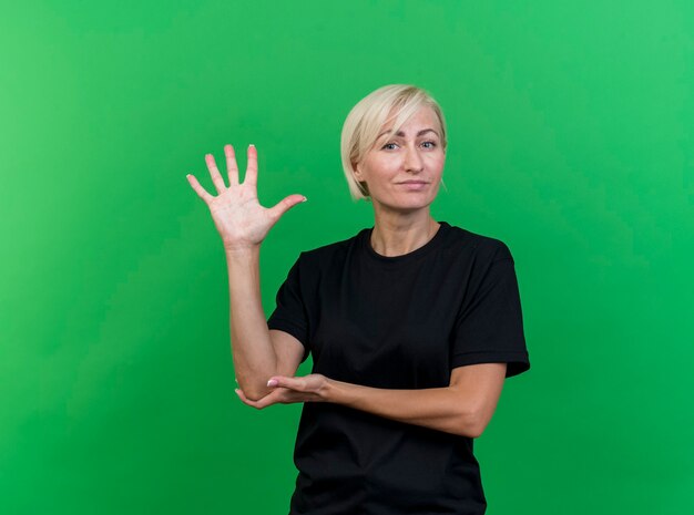 Segura mujer eslava rubia de mediana edad poniendo la mano en el codo mostrando cinco con la mano aislada en la pared verde con espacio de copia
