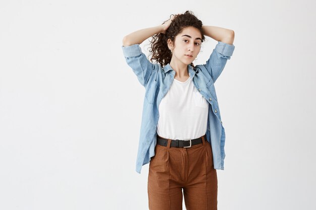 Segura mujer con expresión seria, con camisa vaquera y pantalones marrones, mira a un lado mientras espera algo, usa aretes, juega con el pelo largo y rizado