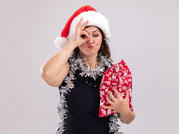 Segura mujer de mediana edad con gorro de Papá Noel y guirnalda de oropel alrededor del cuello sosteniendo un saco de regalo de Navidad mirando a la cámara haciendo gesto de mirada aislado sobre fondo blanco.