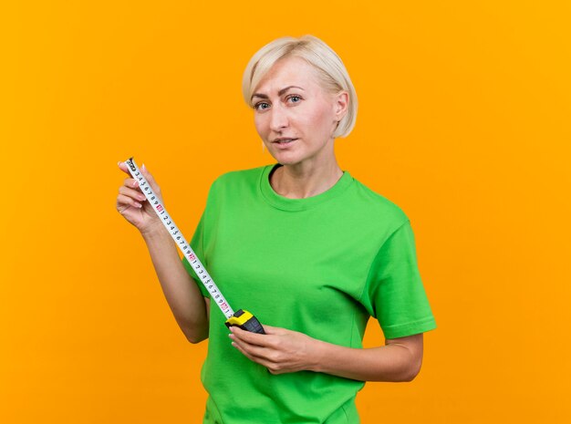 Segura mujer rubia de mediana edad mirando al frente sosteniendo medidor de cinta aislado en la pared amarilla