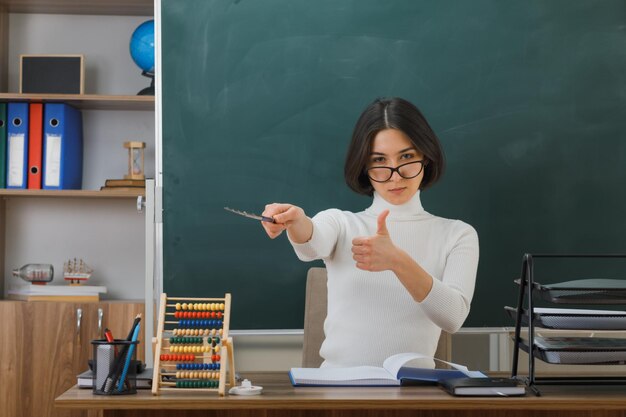 segura de sí misma mostrando los pulgares hacia arriba, una joven maestra que usa anteojos apunta a un lado con un puntero sentado en el escritorio con herramientas escolares en el aula