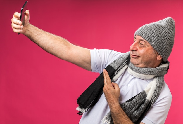 Seguro adulto enfermo hombre caucásico con bufanda alrededor del cuello con gorro de invierno tomando selfie gesticulando signo de victoria aislado en la pared rosa con espacio de copia