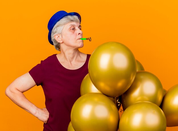 Seguro de anciana con sombrero de fiesta se encuentra con globos de helio que soplan silbato aislado en la pared naranja con espacio de copia