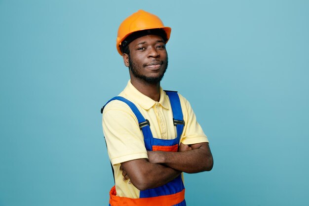 Seguro cruzando las manos joven constructor afroamericano en uniforme aislado sobre fondo azul.