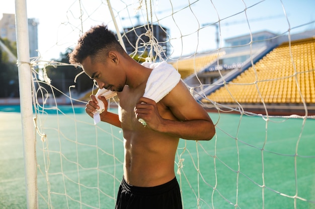 Seguro deportista afroamericano sin camisa con toalla en los hombros después del partido de fútbol en el estadio de la ciudad