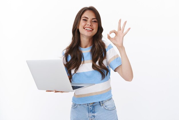 Seguro despreocupado bastante joven sosteniendo la computadora portátil, asegurándose de que ella es hábil y profesional, sonriente, muestra el signo de ok ok