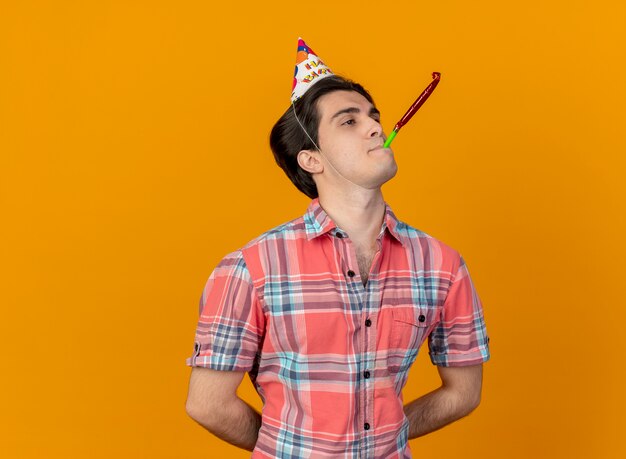 Seguro guapo caucásico hombre vestido con gorro de cumpleaños soplando silbato de fiesta mirando al lado