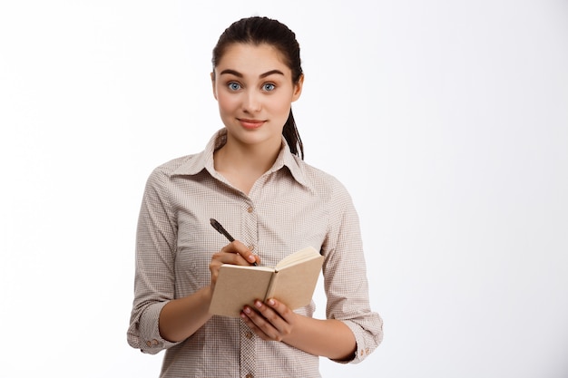 Seguro hermosa joven empresaria escribiendo en cuaderno sonriendo sobre pared blanca