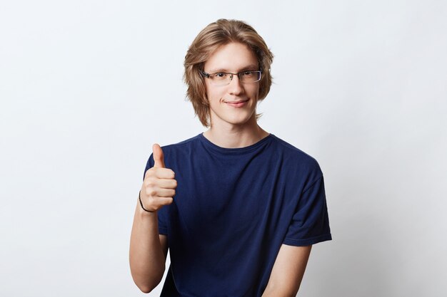 Seguro independiente masculino con elegante peinado, con gafas, mostrando un signo bien mientras levanta el pulgar, de acuerdo con algo. Joven gerente posando en blanco. Concepto de carrera