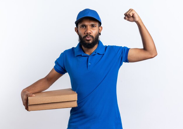 Seguro joven afroamericano repartidor sosteniendo cajas de pizza y levantando el puño aislado sobre fondo blanco con espacio de copia