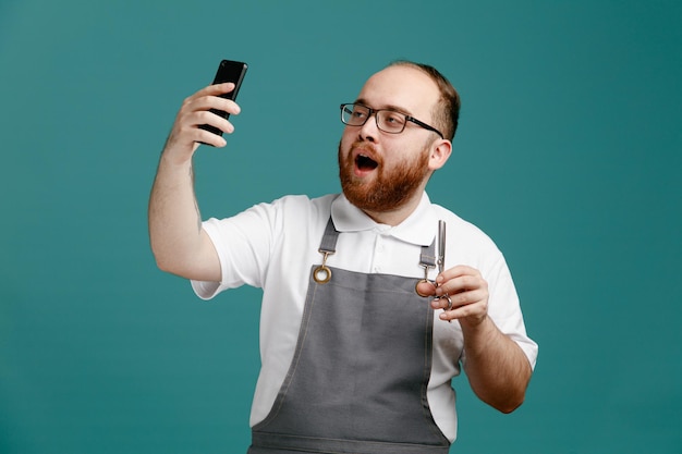 Seguro joven barbero vistiendo uniforme y gafas sosteniendo tijeras tomando selfie con teléfono móvil aislado sobre fondo azul.