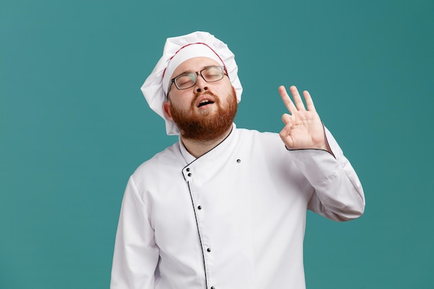 Foto gratuita seguro joven chef masculino con uniforme de gafas y gorra que muestra el signo de ok con los ojos cerrados aislado sobre fondo azul.