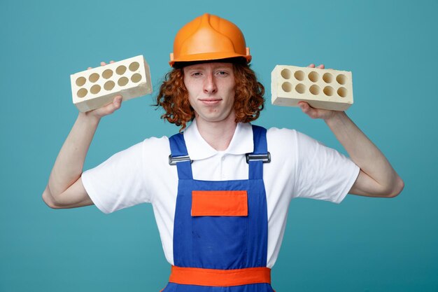 Seguro joven constructor hombre en uniforme sosteniendo ladrillos aislado sobre fondo azul.