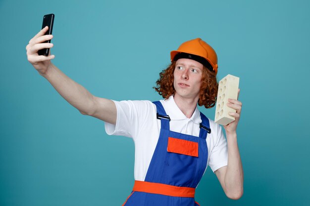 Seguro joven constructor hombre en uniforme tome un selfie sosteniendo ladrillo aislado sobre fondo azul.