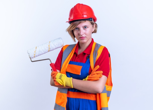 Seguro joven constructor mujer en uniforme con guantes sosteniendo cepillo de rodillo cruzando las manos aisladas en la pared blanca