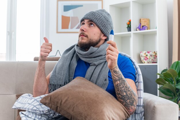 Seguro joven enfermo con bufanda y gorro de invierno sentado en el sofá en la sala de estar con la almohada en las piernas sosteniendo el termómetro mirando hacia arriba mostrando el pulgar hacia arriba