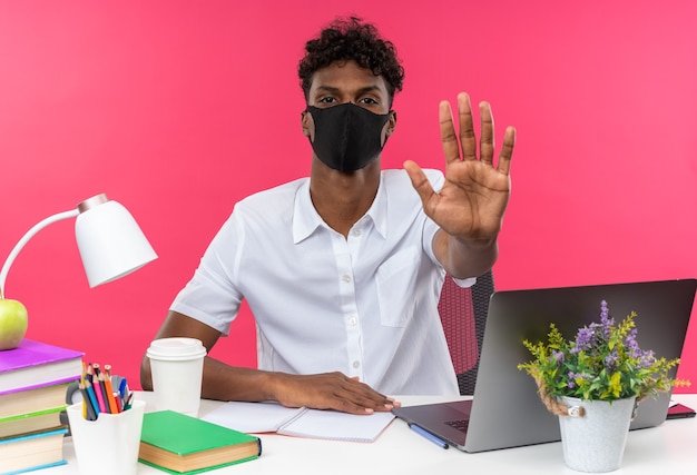 Seguro joven estudiante afroamericano con mascarilla sentado en el escritorio con herramientas escolares gesticulando señal de stop aislada en la pared rosa