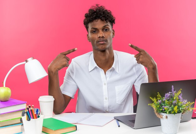 Seguro joven estudiante afroamericano sentado en el escritorio con herramientas escolares apuntando a sí mismo