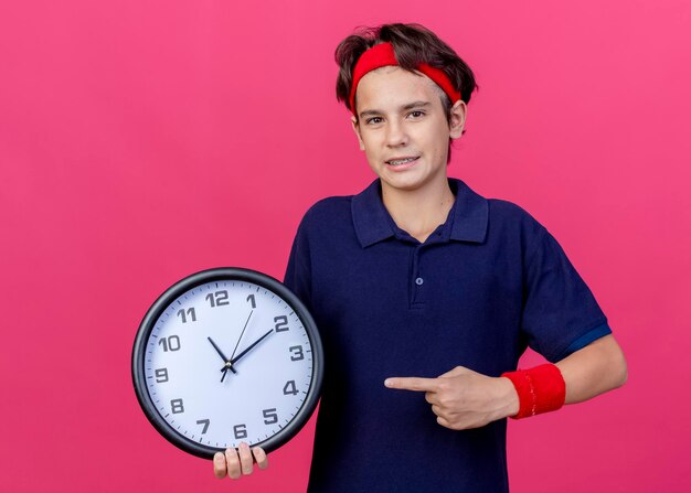 Seguro joven guapo deportivo vistiendo diadema y muñequeras con aparatos dentales sosteniendo y apuntando al reloj mirando al frente aislado en la pared rosa