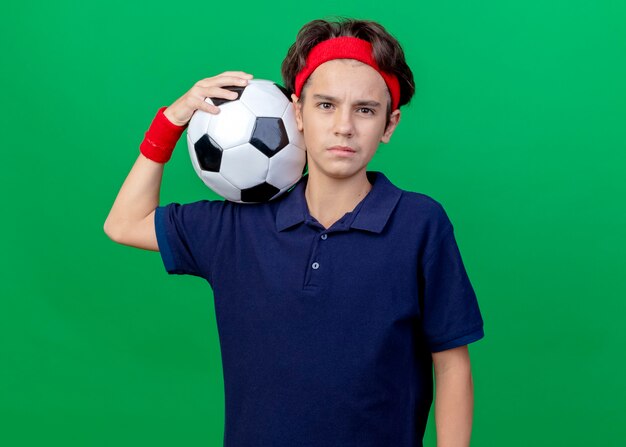 Seguro joven guapo deportivo vistiendo diadema y muñequeras con aparatos dentales sosteniendo un balón de fútbol en el hombro mirando al frente aislado en la pared verde