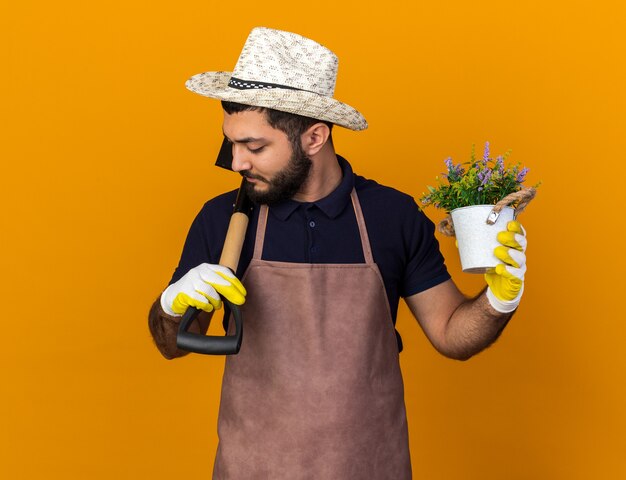 Foto gratuita seguro joven jardinero varón caucásico vistiendo gorro y guantes de jardinería sosteniendo maceta y pala en el hombro aislado en la pared naranja con espacio de copia
