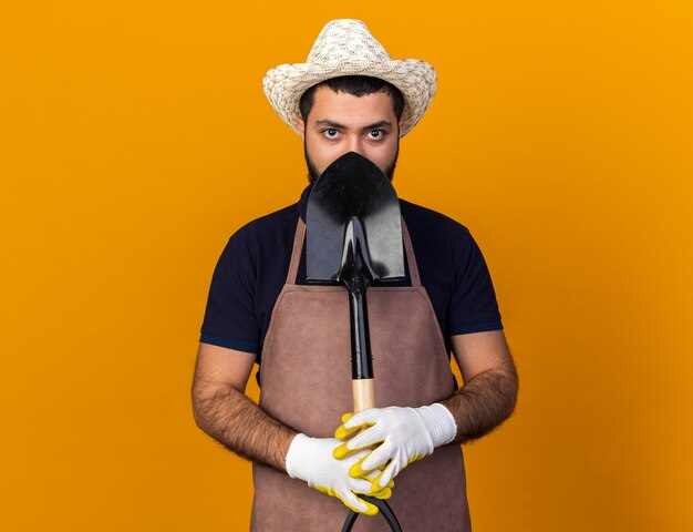 Seguro joven jardinero varón caucásico vistiendo gorro y guantes de jardinería sosteniendo la pala aislado en la pared naranja con espacio de copia
