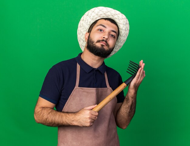 Seguro joven jardinero varón caucásico vistiendo sombrero de jardinería sosteniendo el rastrillo y mirando aislado en la pared verde con espacio de copia