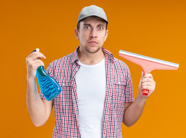 Seguro joven limpiador con gorra sosteniendo agente de limpieza con cabeza de trapeador aislado en la pared naranja