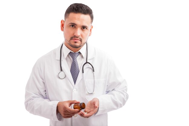 Seguro joven médico hispano tomando algunas pastillas recetadas de una botella en un fondo blanco.