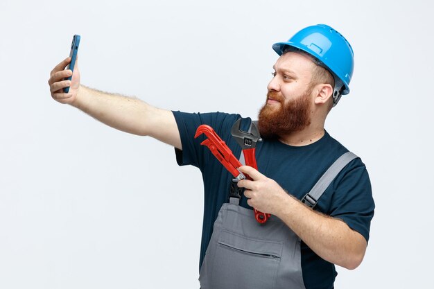 Seguro joven trabajador de la construcción masculino con casco de seguridad y uniforme con llave de tubo y llave inglesa tomando selfie con teléfono móvil aislado sobre fondo blanco.