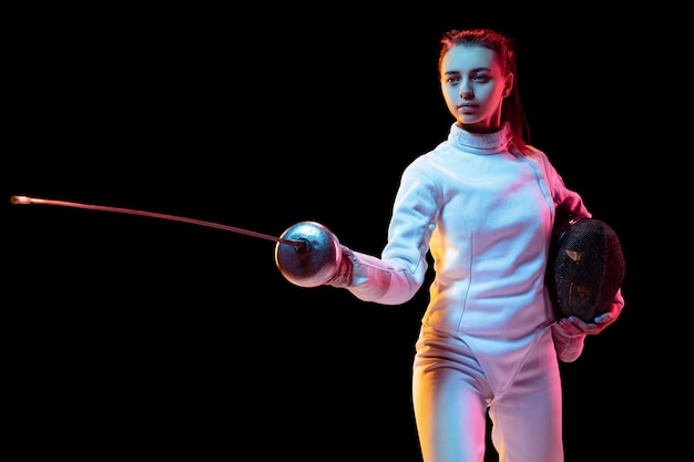 Seguro. Jovencita en traje de esgrima con espada en mano aislada sobre fondo negro, luz de neón. Joven modelo practicando y entrenando en movimiento, acción. Copyspace. Deporte, juventud, estilo de vida saludable.