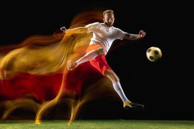 Seguro. Jugador de fútbol o fútbol masculino caucásico joven en ropa deportiva y botas pateando la pelota para el gol en luz mixta en la pared oscura. Concepto de estilo de vida saludable, deporte profesional, afición.