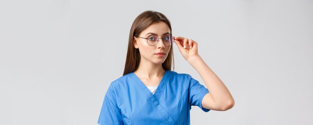 Seguro de medicina para trabajadores de la salud y concepto de pandemia de covid19 Enfermera profesional inteligente o pasante con batas azules y anteojos mirando a la cámara con fondo gris determinado