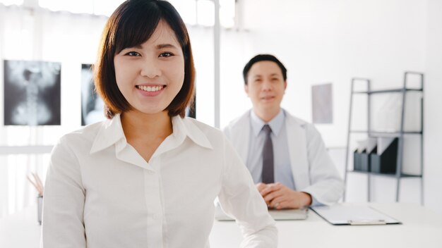 Seguro médico de Asia en uniforme médico blanco y joven paciente mirando a cámara y sonriendo mientras consulta médica en el escritorio en la clínica de salud u hospital.