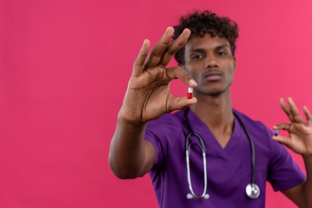 Un seguro médico joven guapo de piel oscura con cabello rizado vistiendo uniforme violeta con estetoscopio mirando pastillas