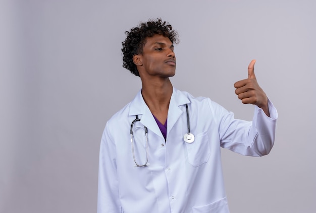 Un seguro médico joven guapo de piel oscura con pelo rizado vistiendo bata blanca con estetoscopio mostrando los pulgares para arriba