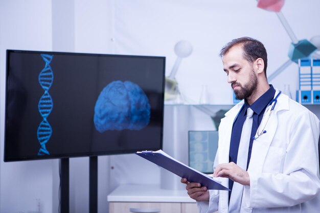 Seguro médico joven con un portapapeles en sus manos de pie junto a un monitor con un gráfico cerebral. Doctor guapo.