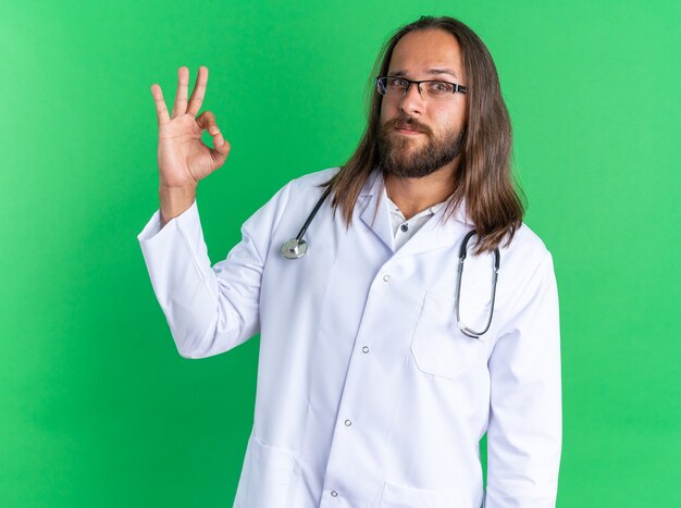 Seguro médico varón adulto vistiendo bata médica y un estetoscopio con gafas mirando a la cámara haciendo el signo de ok aislado en la pared verde