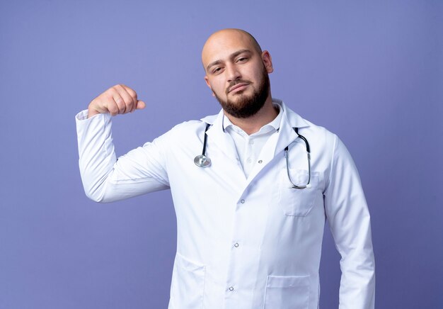 Seguro médico varón joven calvo vistiendo bata médica y estetoscopio haciendo gesto fuerte aislado sobre fondo azul.