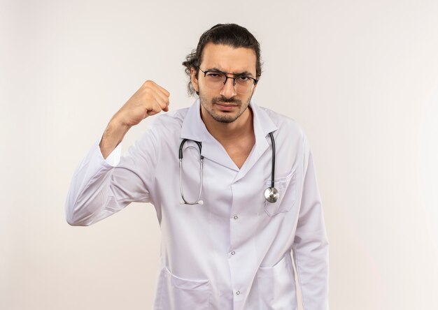 Seguro médico varón joven con gafas ópticas vistiendo túnica blanca con estetoscopio haciendo gesto fuerte