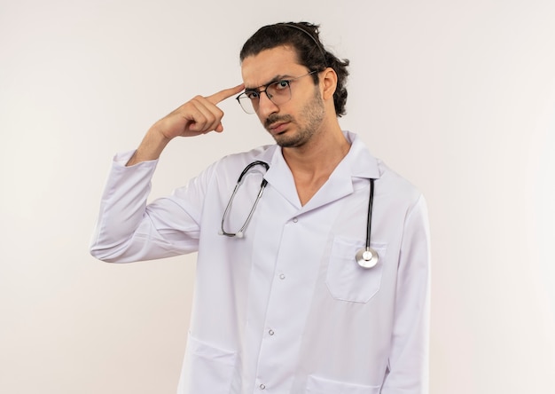 Seguro médico varón joven con gafas ópticas vistiendo túnica blanca con estetoscopio poniendo el dedo en la frente