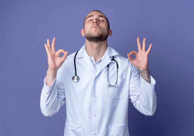 Seguro médico varón joven vistiendo bata médica y un estetoscopio haciendo bien los signos con los ojos cerrados aislados en púrpura con espacio de copia