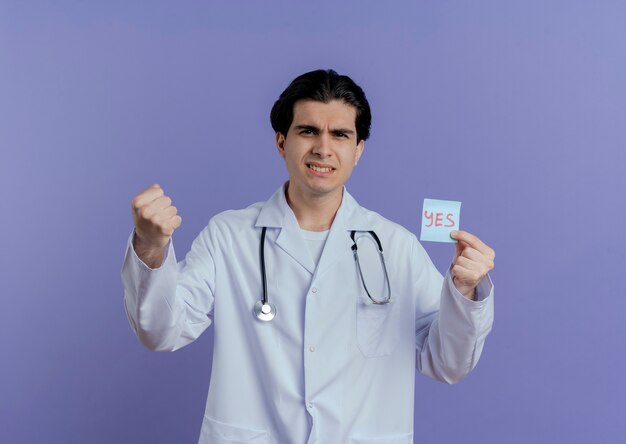 Seguro médico varón joven vistiendo bata médica y un estetoscopio mostrando sí nota haciendo gesto fuerte aislado en la pared púrpura con espacio de copia