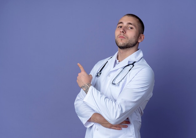 Seguro médico varón joven vistiendo bata médica y estetoscopio de pie en la vista de perfil poniendo la mano sobre el riñón y apuntando al lado aislado en púrpura con espacio de copia