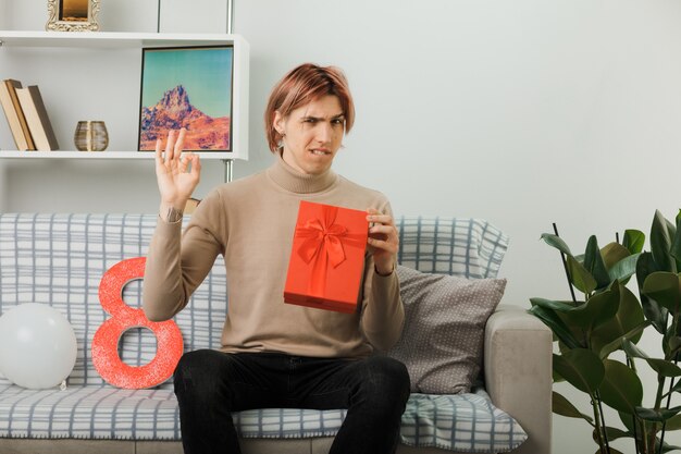 Seguro mostrando buen gesto chico guapo en el día de la mujer feliz celebración presente sentado en el sofá en la sala de estar