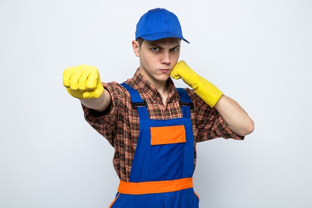 Seguro de pie en pose de lucha joven chico de limpieza con uniforme y gorra con guantes