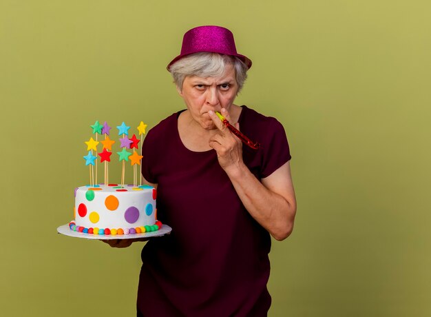 Seguros de anciana vistiendo gorro de fiesta soplando silbato sosteniendo pastel de cumpleaños aislado en la pared verde oliva con espacio de copia
