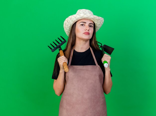 Seguros de hermosa chica jardinero en uniforme con sombrero de jardinería poniendo rastrillo con rastrillo de azada en el hombro aislado en verde